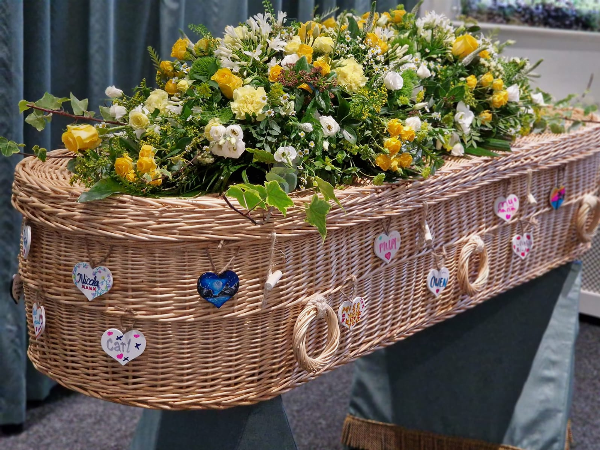 Light Willow Wicker Coffin Available at Jason O'Brien & Family Funeral Services Limited in Ashton-Under-Lyne, Greater Manchester. Adorned with a yellow and white mixed floral arrangement and personalised with painted hearts supplied by family and friends.